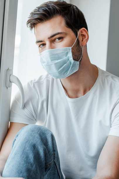 Hombre con máscara médica mirando a la cámara cerca de la ventana
  - Foto, imagen