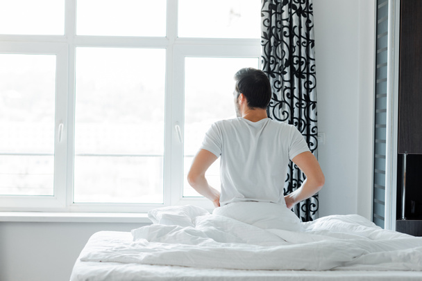 Back view of man looking at window while sitting on bed at home   - Photo, Image