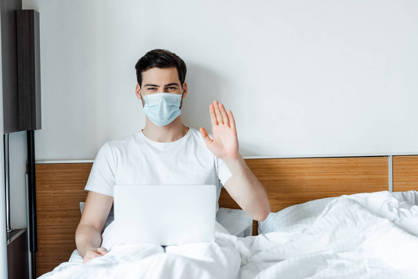 Hombre con máscara médica saludando la mano a la cámara mientras está sentado con el ordenador portátil en la cama
  - Foto, Imagen
