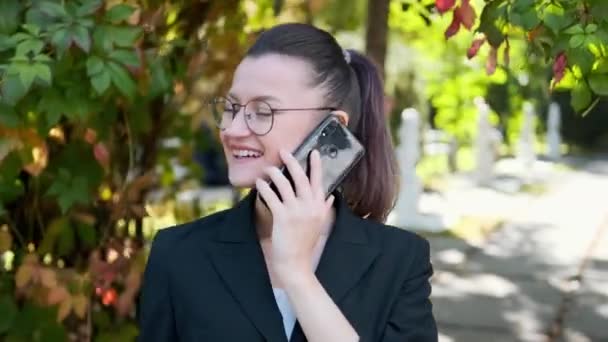 Attractive young business woman with briefcase in black jacket and glasses steps, walks and dances outdoors. Speaks on phone and laughs. Close-up of legs and head. - 映像、動画