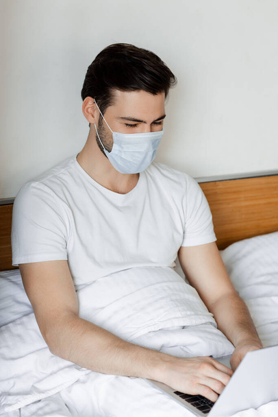 Freelancer in medical mask working with laptop in bedroom - Photo, Image