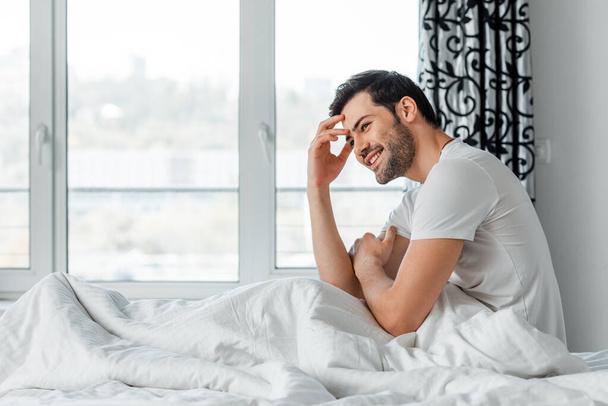 Side view of handsome man smiling away while sitting on bed  - Fotó, kép