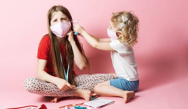 Personal protective equipment: masked sisters play d doctor, and the younger sister treats the older and looks into her ear. Portrait on a pink background. - Photo, Image