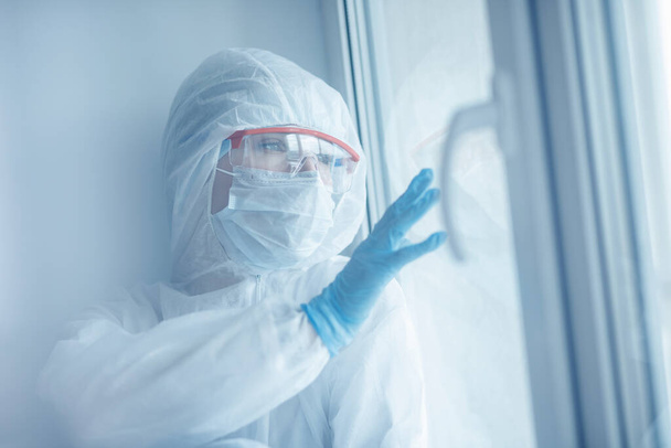 Loneliness quarantine, young woman in medical mask looks out window self isolation. Coronavirus pandemic concept - Φωτογραφία, εικόνα