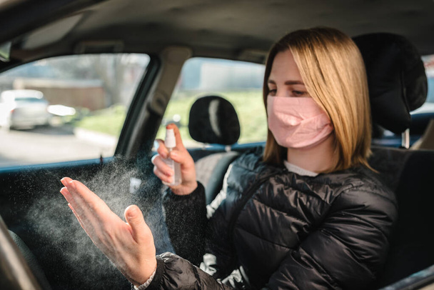 Spraying antibacterial sanitizer spray on hand in car, infection control concept. Sanitizer to prevent Coronavirus, COVID-19, flu. Spray bottle. Woman wearing in medical protective mask driving a car. - Fotoğraf, Görsel