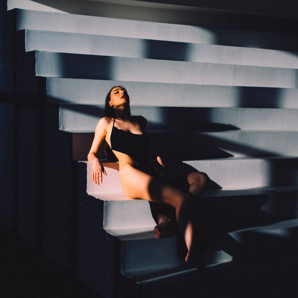 In the evening, a young beautiful girl is alone in an empty studio, she has almost no clothes on her, she is naked, the setting sun through the windows falls on her body and gives off Shadows and contours - Photo, image