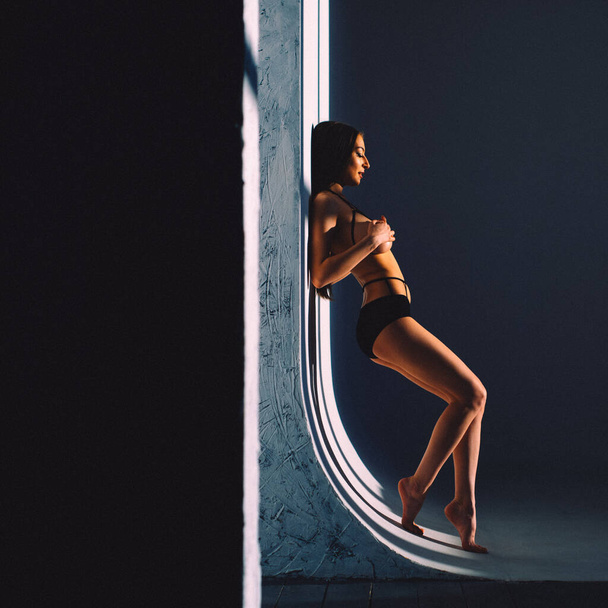 In the evening, a young beautiful girl is alone in an empty studio, she has almost no clothes on her, she is naked, the setting sun through the windows falls on her body and gives off Shadows and contours - Foto, afbeelding