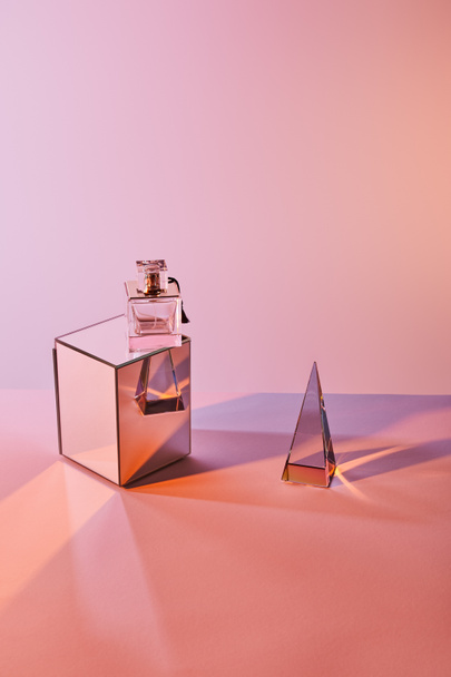 crystal transparent pyramid near perfume bottle on cube on pink background - Foto, imagen