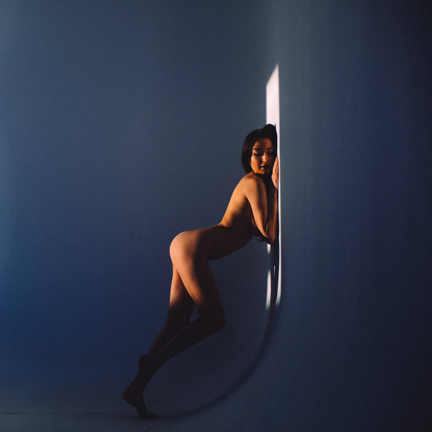 In the evening, a young beautiful girl is alone in an empty studio, she has almost no clothes on her, she is naked, the setting sun through the windows falls on her body and gives off Shadows and contours - Foto, imagen