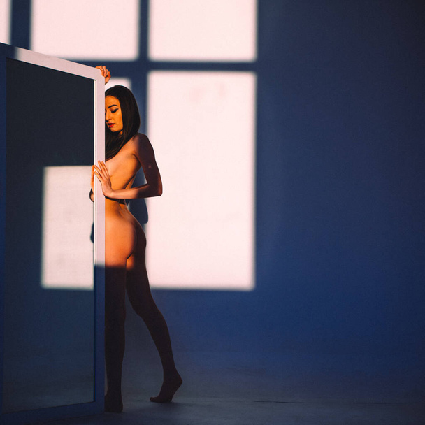 In the evening, a young beautiful girl is alone in an empty studio, she has almost no clothes on her, she is naked, the setting sun through the windows falls on her body and gives off Shadows and contours - Foto, imagen