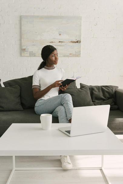thoughtful african american freelancer holding notebook while sitting on sofa near table with laptop - 写真・画像