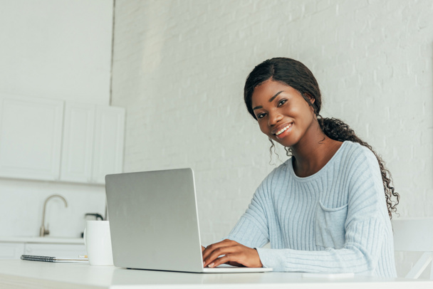 cheerful african american freelancer smiling at camera while working on laptop in kitchen - Фото, изображение