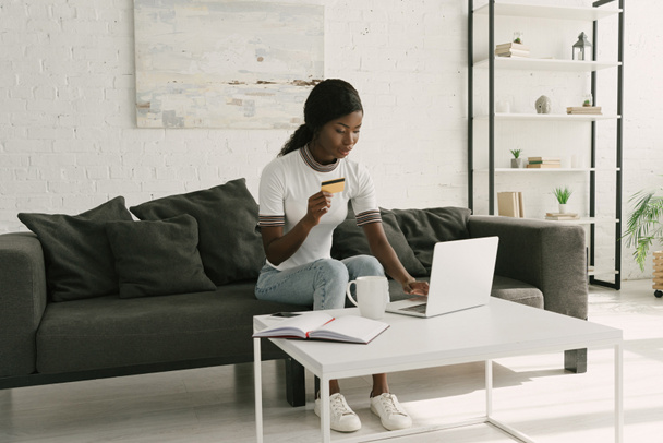 pretty african american girl using laptop and holding credit card while sitting on sofa at home - Zdjęcie, obraz