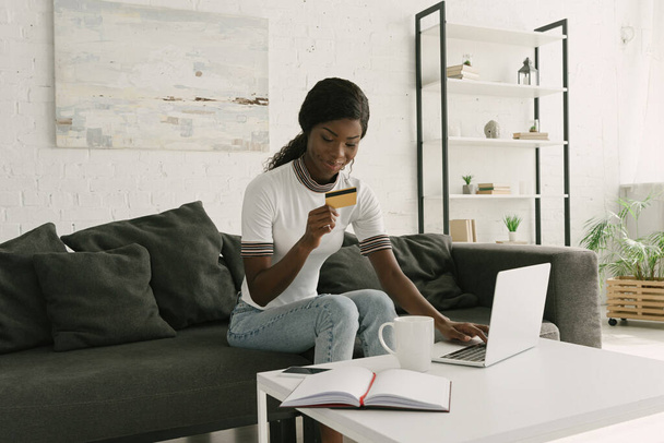 усміхнена афроамериканська дівчина тримає кредитну картку, сидячи біля столу з ноутбуком, ноутбуком та чашкою кави
 - Фото, зображення
