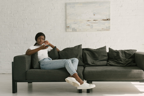 ελκυστική Αφροαμερικανή κοπέλα κρατώντας τηλεχειριστήριο τηλεόρασης, ενώ κάθεται στον καναπέ στο σπίτι - Φωτογραφία, εικόνα