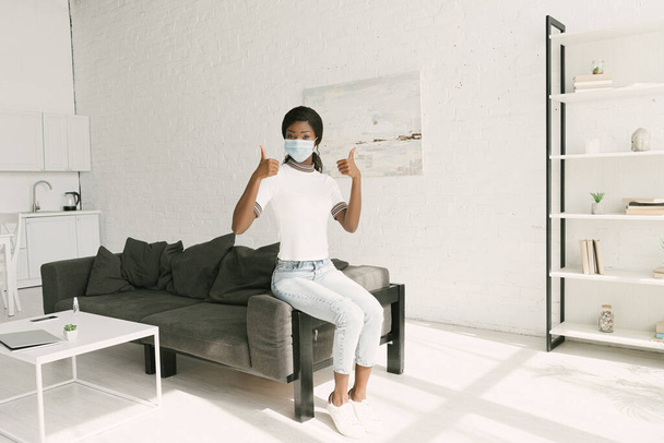 junge afrikanisch-amerikanische Mädchen in medizinischer Maske zeigt Daumen nach oben, während sie auf grauem Sofa in der Küche sitzt - Foto, Bild