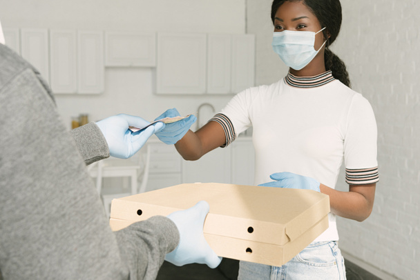 africano americano chica en médico máscara dando dinero a comida entrega hombre y tomando pizza cajas
 - Foto, imagen