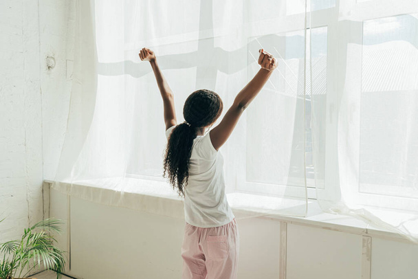 Rückansicht eines afrikanisch-amerikanischen Mädchens im Pyjama, das die Hände ausstreckt, während es im Sonnenlicht am Fenster steht - Foto, Bild
