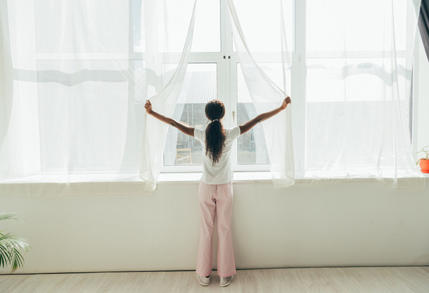 Rückansicht eines afrikanisch-amerikanischen Mädchens im Pyjama, das Fenstervorhänge im Sonnenlicht öffnet - Foto, Bild