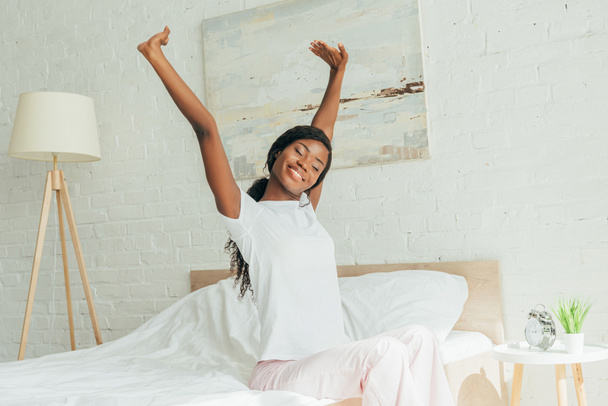 χαμογελαστή Αφροαμερικανίδα με πιτζάμες που κάθεται στο κρεβάτι με κλειστά μάτια και τεντώνεται με σηκωμένα χέρια - Φωτογραφία, εικόνα