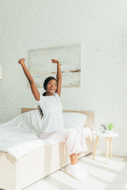 glückliches afrikanisches amerikanisches Mädchen im Pyjama, das sich im Sitzen auf dem Bett mit erhobenen Händen dehnt - Foto, Bild