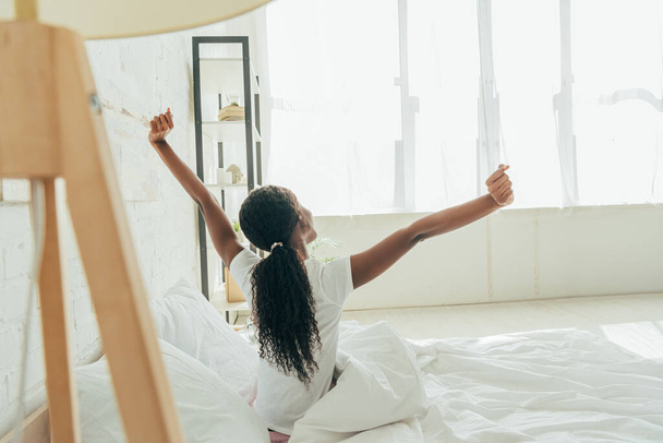 hátsó kilátás afro-amerikai lány nyújtózkodik emelt kézzel, miközben ül az ágyban - Fotó, kép