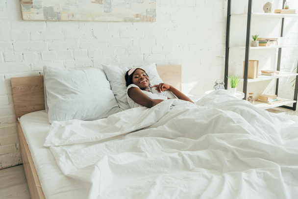 Χαρούμενη Αφροαμερικανή με μάσκα ύπνου στο μέτωπο χαμογελώντας ξαπλωμένη στο κρεβάτι - Φωτογραφία, εικόνα