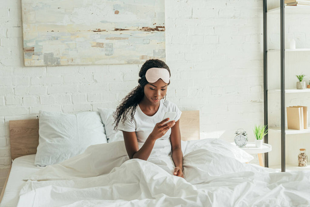 hübsche Afroamerikanerin mit Schlafmaske auf der Stirn plaudert auf Smarthone im Bett - Foto, Bild