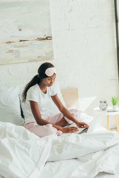 young african american freelancer with sleep mask on forehead using laptop in bed - Photo, Image