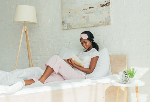 attentif afro-américaine fille avec masque de sommeil sur le front livre de lecture tout en étant assis dans le lit
 - Photo, image