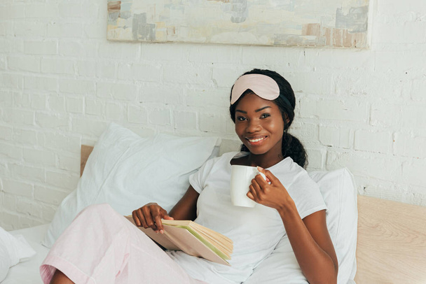 chica afroamericana feliz con máscara del sueño en la frente sentado en la cama, sosteniendo libro y taza de café
 - Foto, imagen