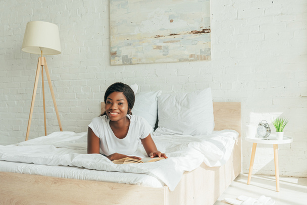cheerful african american girl lying on bed with book and smiling at camera - Фото, зображення
