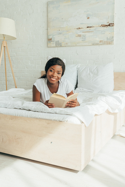 glückliches afrikanisch-amerikanisches Mädchen liegt mit offenem Buch auf weißem Bettzeug - Foto, Bild