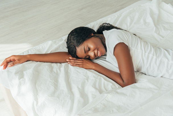 vista de ángulo alto de hermosa, sonriente chica afroamericana acostada sobre ropa de cama blanca con los ojos cerrados
 - Foto, Imagen