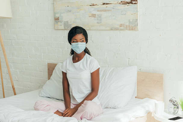 dreamy african american woman in medical mask sitting in bed with crossed legs and looking away - Фото, изображение