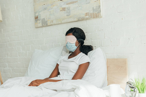 fiatal afro-amerikai nő orvosi maszk és alvás maszk pihen, miközben ül az ágyban - Fotó, kép