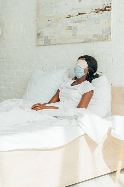 menina afro-americana em máscara médica e máscara de sono descansando enquanto sentado na cama
 - Foto, Imagem