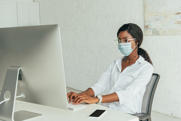 attentive african american freelancer in medical mask typing on keyboard near computer monitor at home - Fotografie, Obrázek