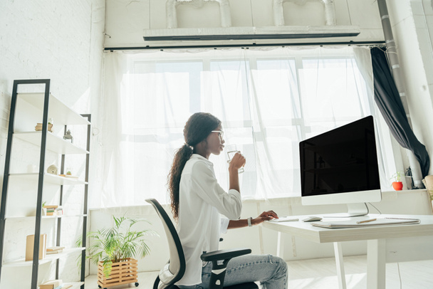 vista lateral del freelancer afroamericano sosteniendo un vaso de agua y mirando el monitor de computadora con pantalla en blanco mientras trabaja en casa
 - Foto, imagen
