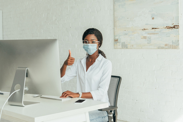 young african american freelancer in medical mask showing thumb up while sitting near computer monitor at home - Photo, Image