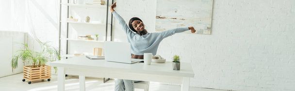 horizontal crop of happy african american freelancer stretching while sitting at table near laptop  - Fotografie, Obrázek