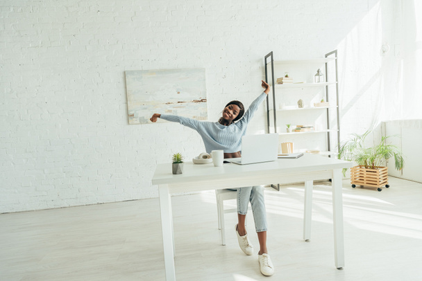 smiling african american freelancer stretching while sitting near laptop in spacious living room - Photo, Image