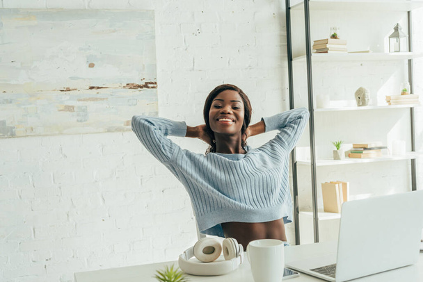 alegre afroamericano freelancer estiramiento mientras está sentado cerca de la computadora portátil, auriculares inalámbricos y taza de café
 - Foto, imagen