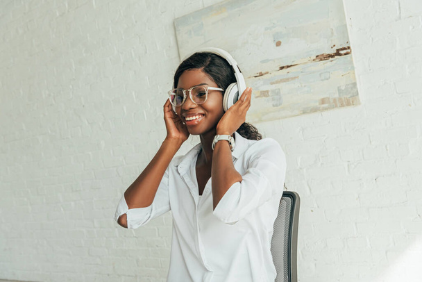 feliz afroamericana chica en gafas tocando auriculares inalámbricos mientras escucha música
 - Foto, Imagen