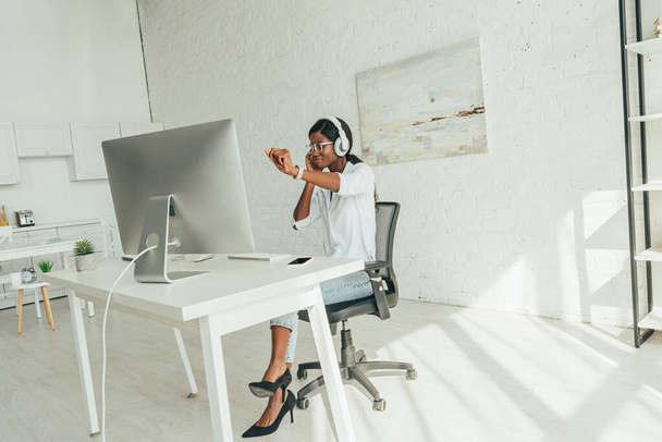 alegre afroamericano freelancer gesto mientras escucha música en auriculares inalámbricos cerca del monitor de la computadora
 - Foto, imagen