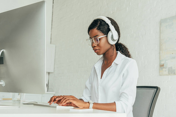 attento freelance afroamericano in cuffie senza fili e occhiali digitando sulla tastiera del computer
 - Foto, immagini