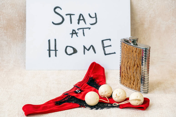 Mutandine da bikini rosse femminili, fiaschetta di metallo con bevanda alcolica, cartello "Stay home" e sex toy di quattro palle vaginali per una soddisfazione sensuale
. - Foto, immagini
