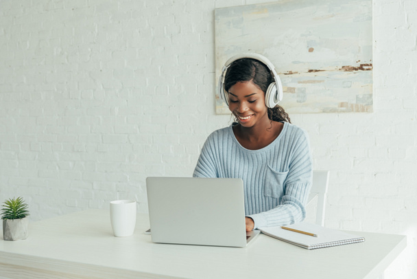 glimlachend Afrikaans Amerikaans freelancer in draadloze hoofdtelefoon werken op laptop thuis - Foto, afbeelding