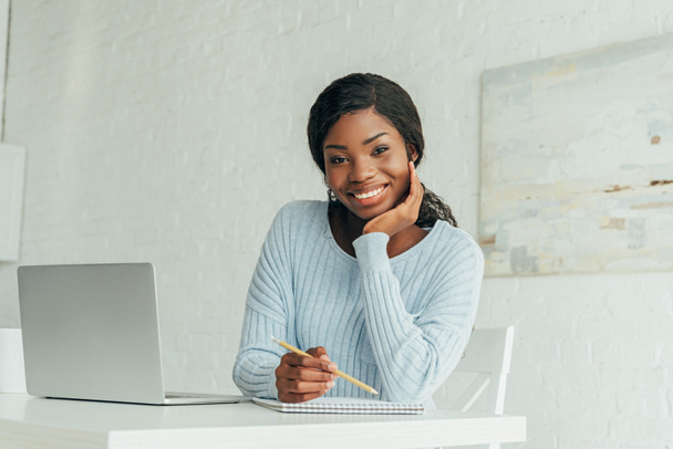 happy african american freelancer smiling at camera while sitting near laptop - Foto, Bild
