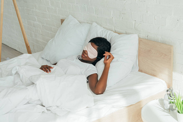 mladá africká americká žena spí na bílém lůžkoviny ve spánkové masce - Fotografie, Obrázek